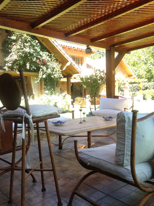 Outdoor patio with wooden pergola, table, chairs with white cushions, and view of a house and garden.