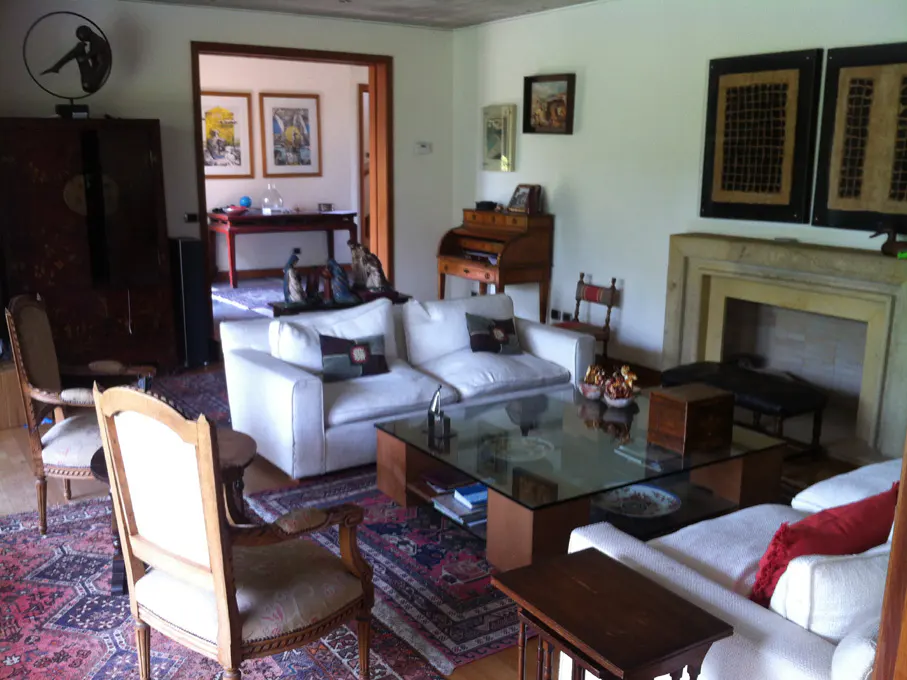 A living room with white sofas, a glass coffee table, and a fireplace. Art hangs on the walls, and a patterned rug covers the floor.