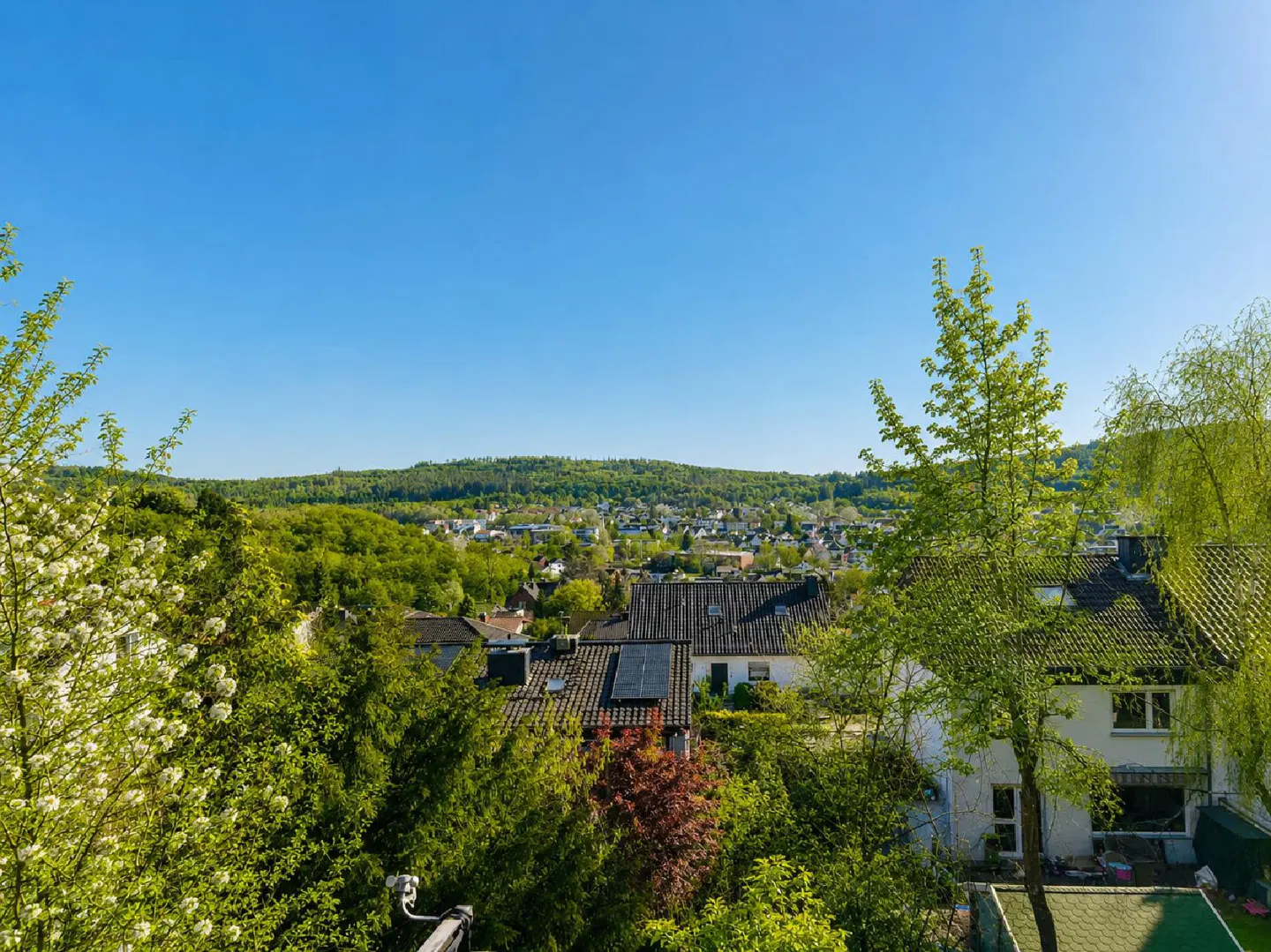 View of a town nestled in green hills under a clear blue sky, seen from a high vantage point. Houses with dark roofs are surrounded by lush trees.