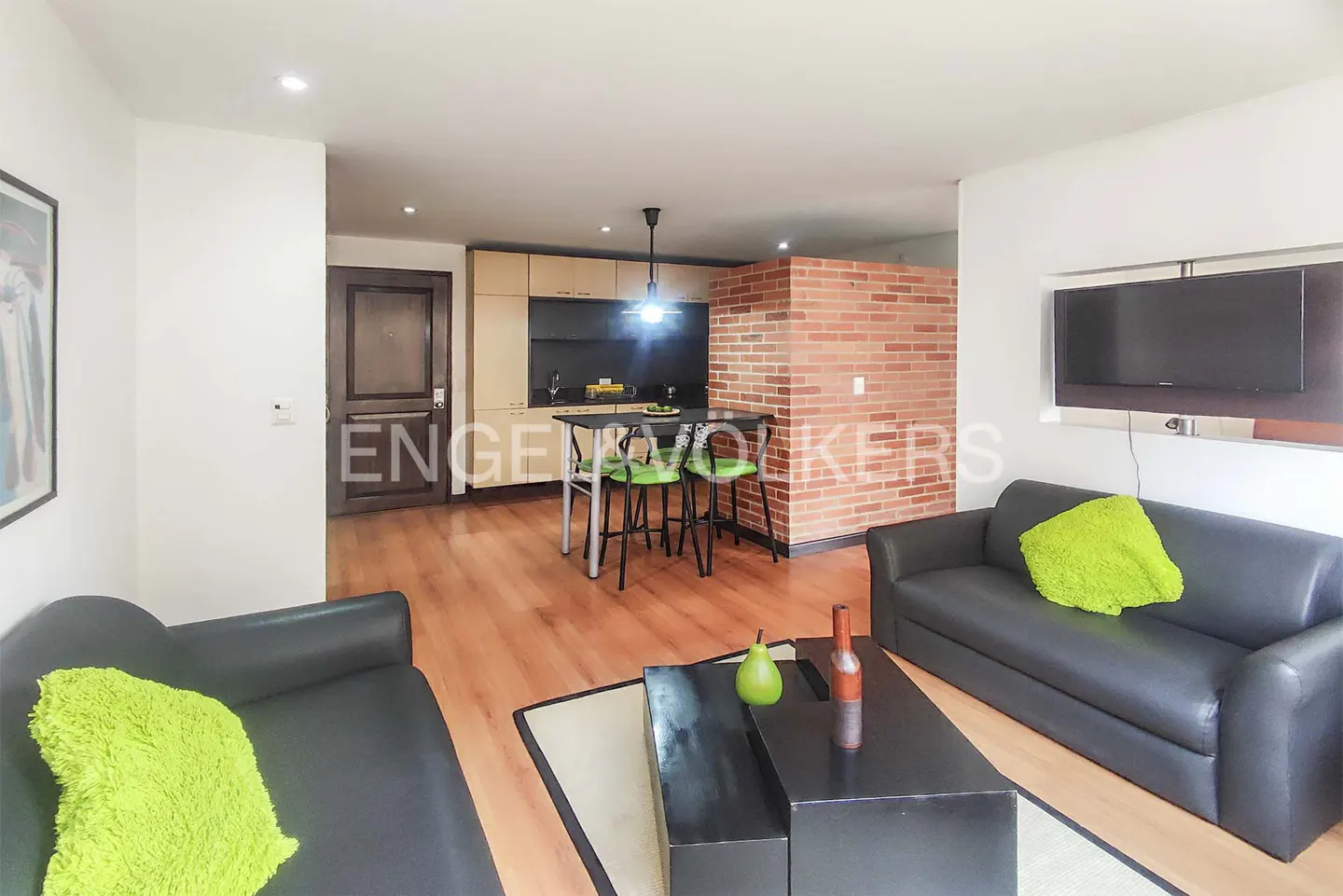 Living room with black sofas, green pillows, and a black coffee table. Kitchen with brick wall and bar stools in the background.