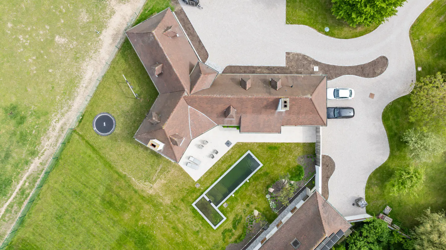 Aerial view of a large house with a brown roof, a pool, a trampoline, and two cars parked in the driveway.