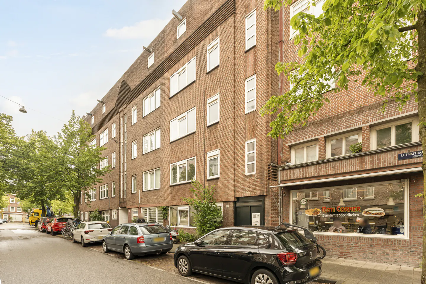 Exterior view of a brick apartment building with a storefront, cars parked on the street, and green trees.