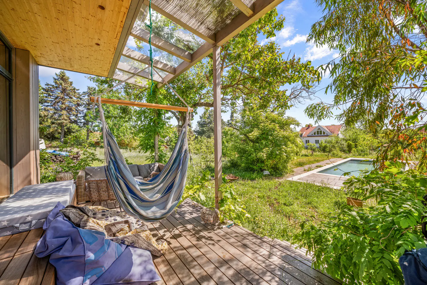 Wooden deck with a hammock, pillows, and a couch. A pool and house are visible in the green backyard.