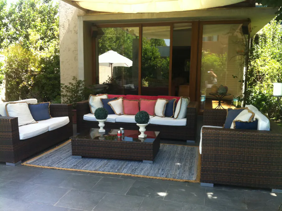 Outdoor patio with brown wicker furniture, white cushions, and colorful pillows. A blue rug sits on the gray stone patio. Trees in the background.