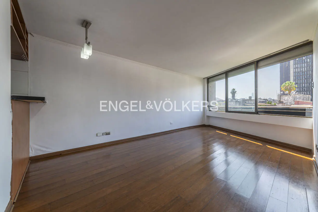 A bright, empty room with hardwood floors and a large window overlooking a city. The walls are white, and the Engel & Völkers logo is visible.