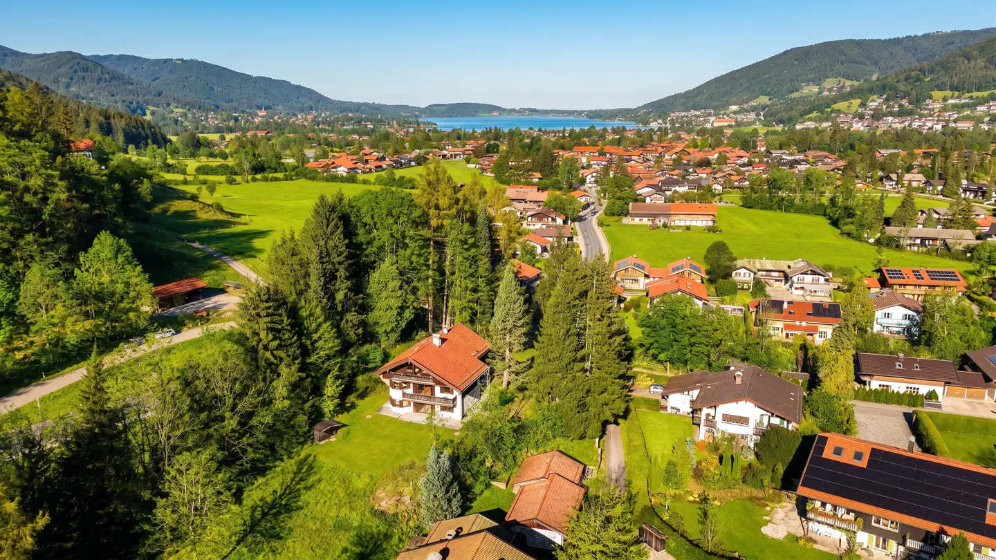 Aerial view of a village nestled in a valley, with houses, trees, and a lake in the background.