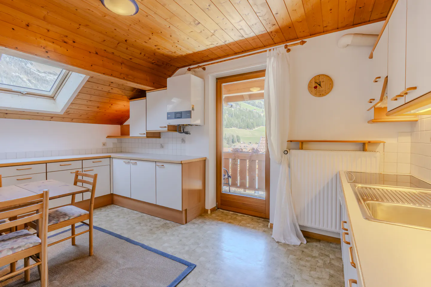 A bright kitchen with wood ceiling, white cabinets, and a door to a balcony with a mountain view. A table and chairs are on a blue rug.