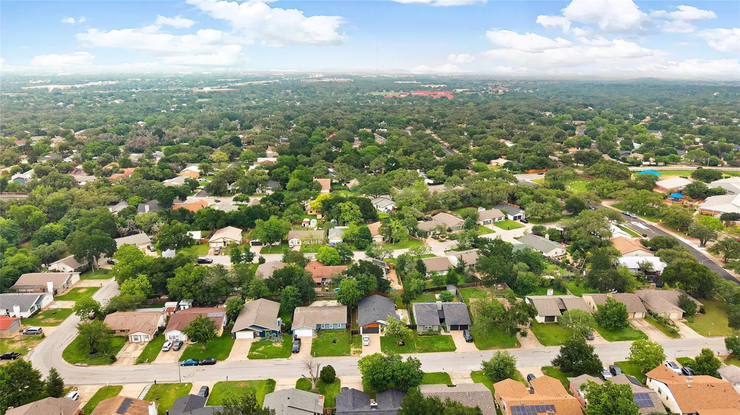 Aerial view of a suburban neighborhood with houses, green trees, and blue sky with clouds.