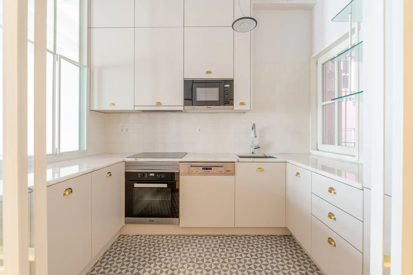 Bright kitchen with white cabinets, gold hardware, and geometric tile floor. Appliances include a microwave, oven, and dishwasher. Natural light from windows.