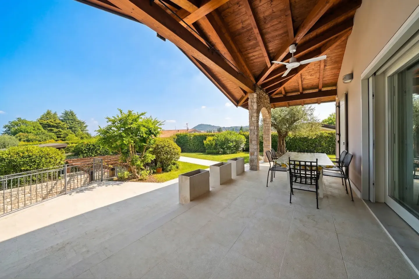 Covered patio with a wood ceiling, stone columns, and a ceiling fan. A table and chairs sit on the patio, overlooking a green lawn and trees.