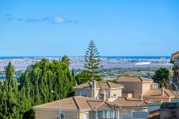 View of beige houses with orange tile roofs, green trees, and a city skyline across a body of water under a blue sky.