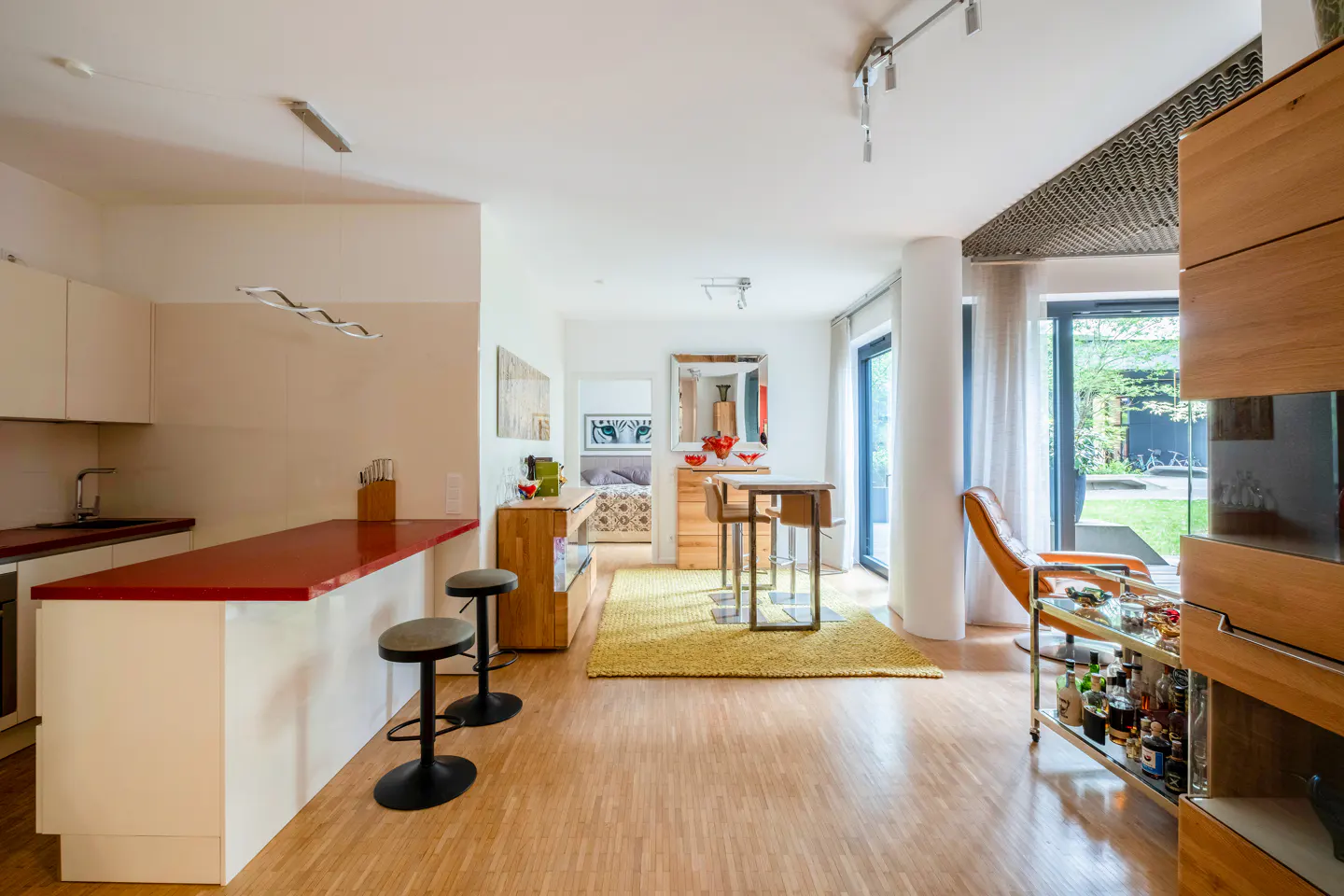 Modern open-plan kitchen with a red breakfast bar, two stools, yellow area rug, dining table, and view of a garden.