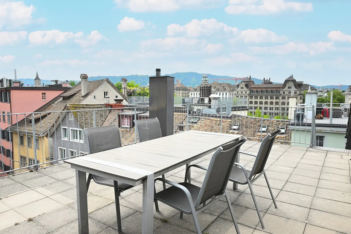 Rooftop patio with a white table and four gray chairs. City buildings and a blue sky with clouds are in the background.
