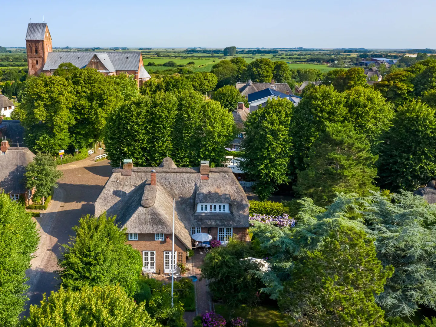 Aerial view of a thatched-roof house surrounded by green trees, with a church in the background on a sunny day.
