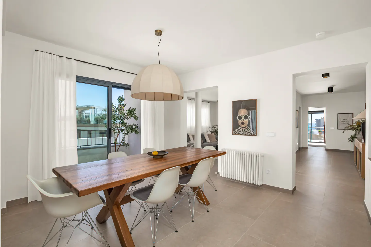 Bright dining room with a wooden table, white chairs, and a large window with sheer curtains. A modern painting hangs on the white wall.