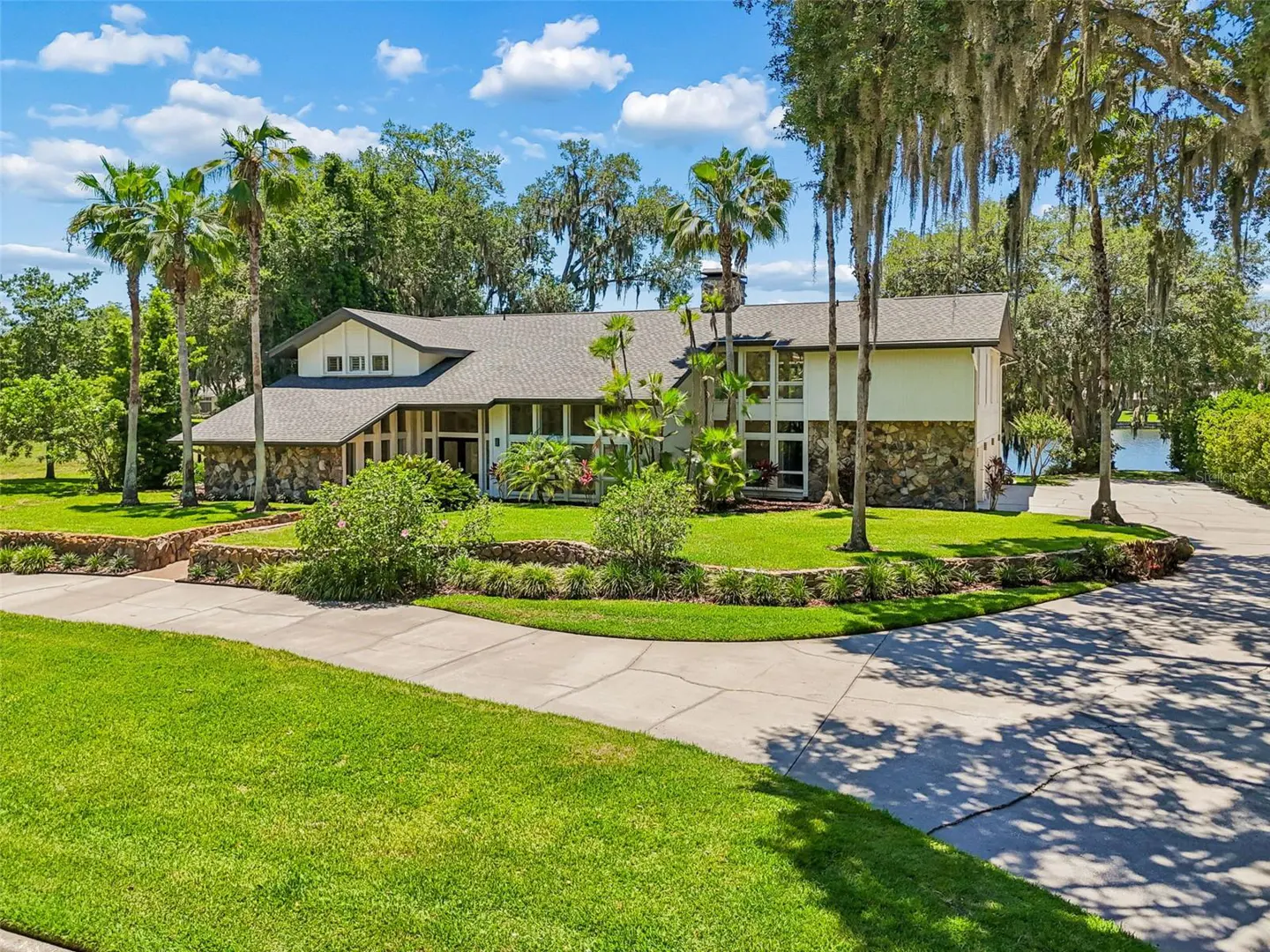 A large, two-story house with a stone facade, gray roof, and palm trees, sits on a green lawn under a blue sky.