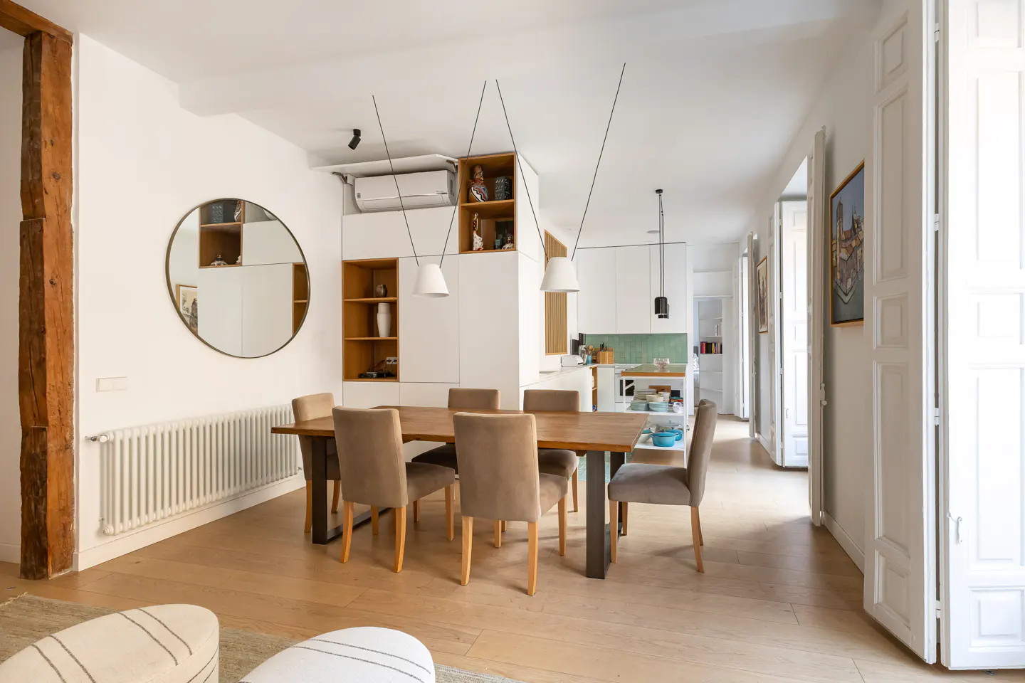 Bright dining room with a wooden table, six taupe chairs, and a round mirror on a white wall. Open kitchen and hallway visible.