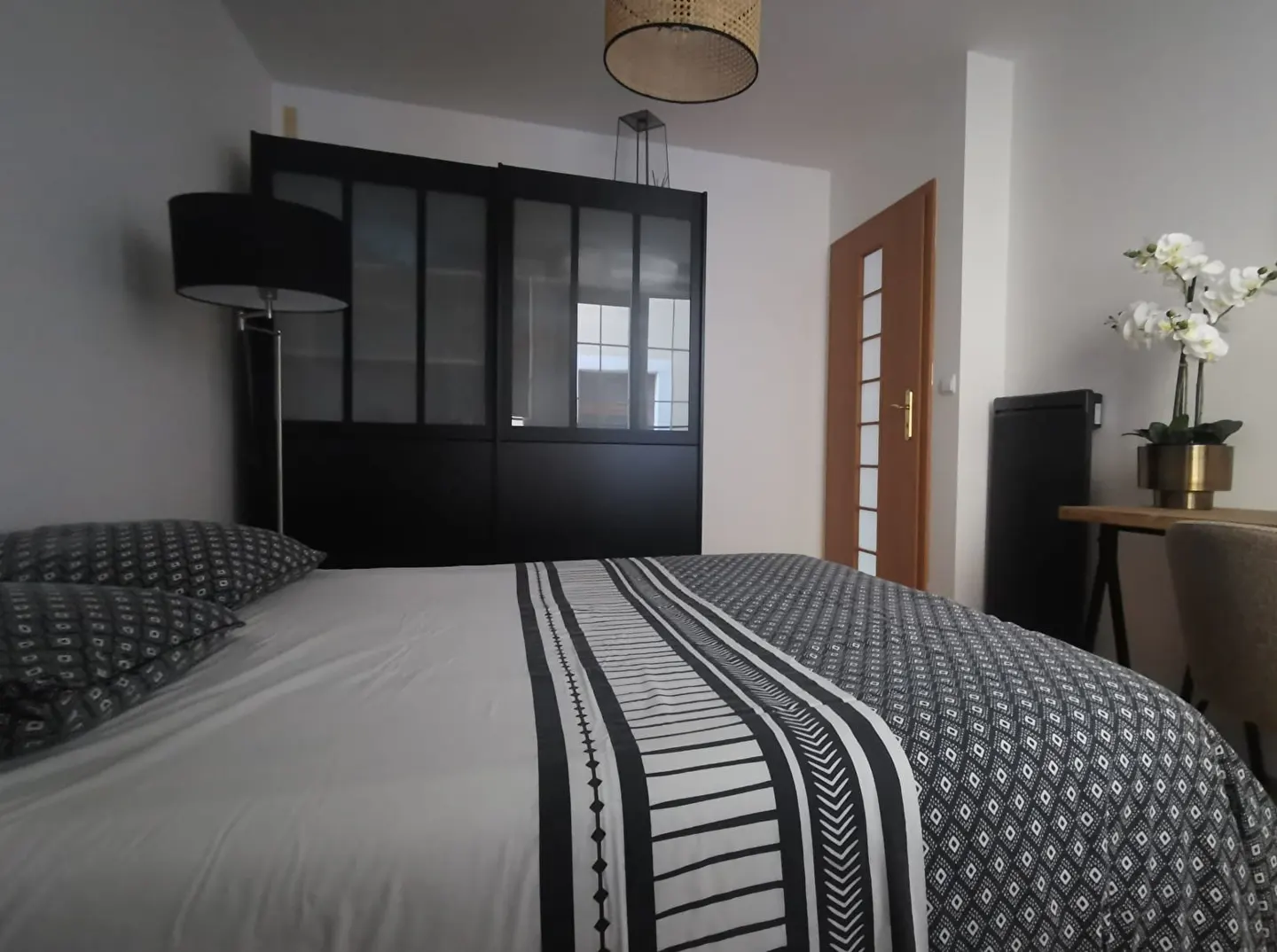 Bedroom with a bed with black and white patterned bedding, a black wardrobe, and a desk with white flowers.
