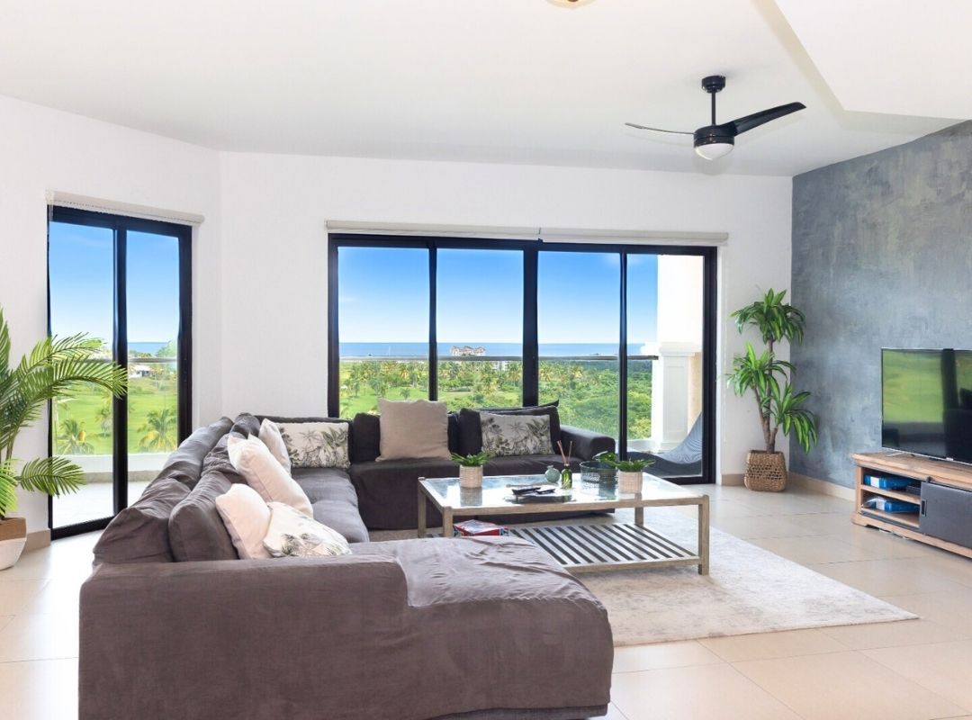 A bright living room with a gray sectional sofa, a glass coffee table, and a view of the ocean through large windows.