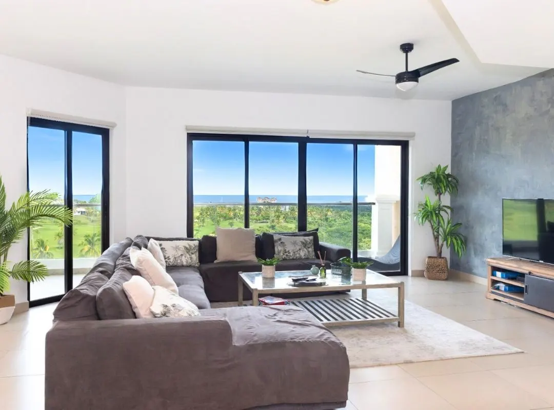 A bright living room with a gray sectional sofa, a glass coffee table, and a view of the ocean through large windows.