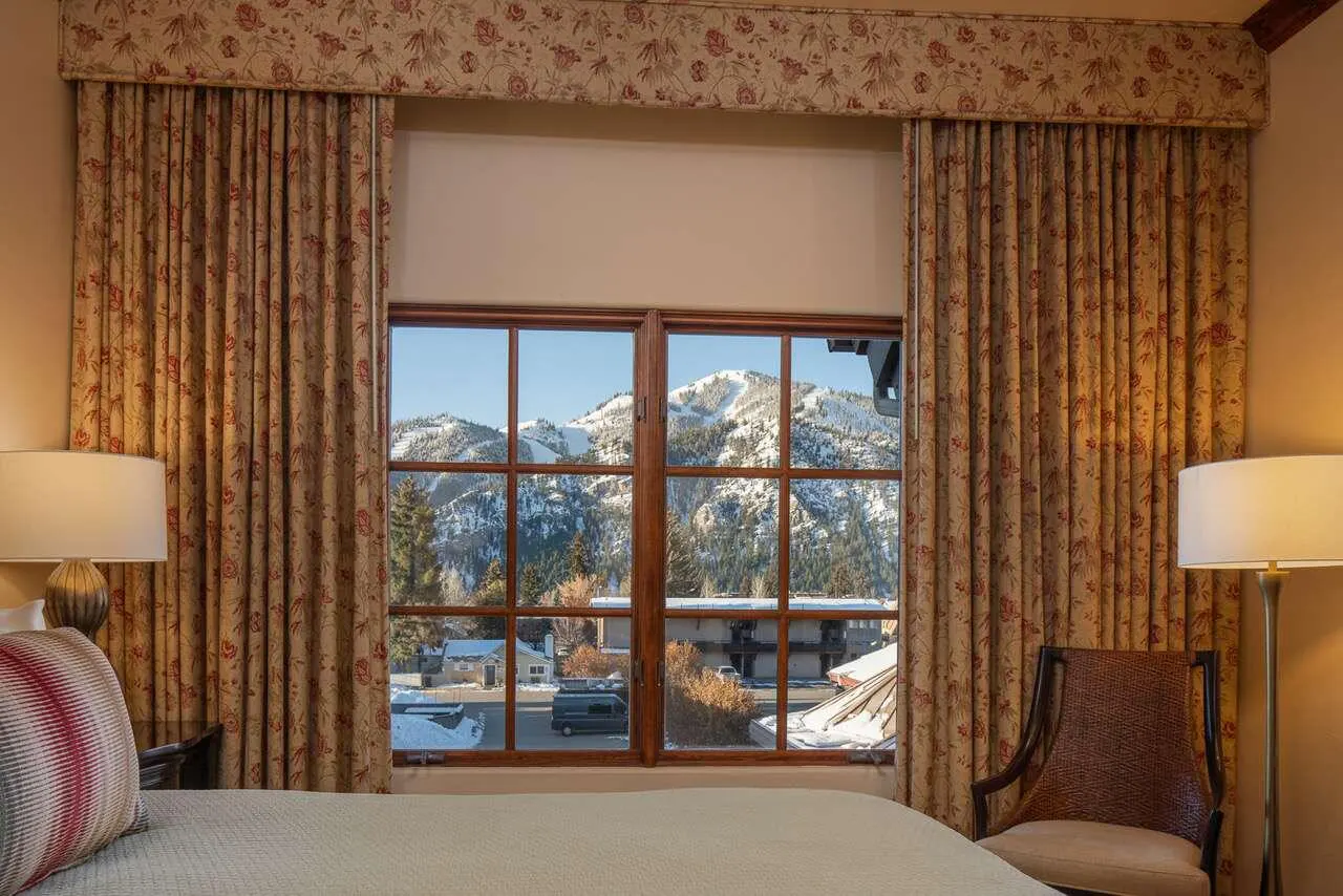 Bedroom view with floral curtains framing a window. Outside, snow-covered mountains and a clear blue sky are visible.