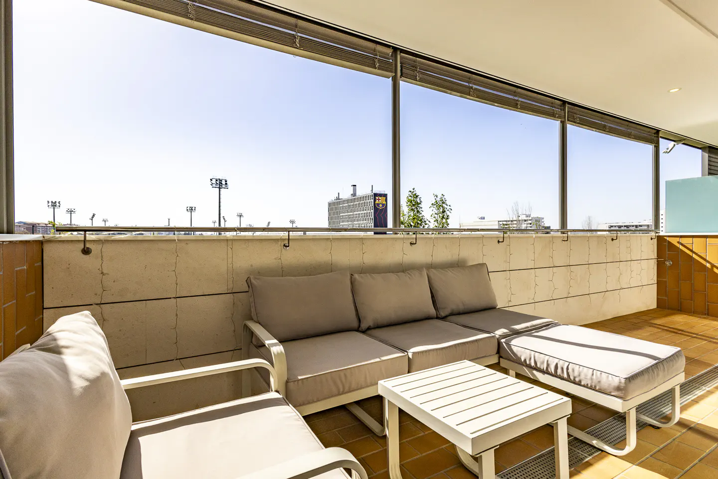 Outdoor patio with a beige sofa, chair, and table on a terracotta tile floor. A building with a blue and gold logo is visible in the background.