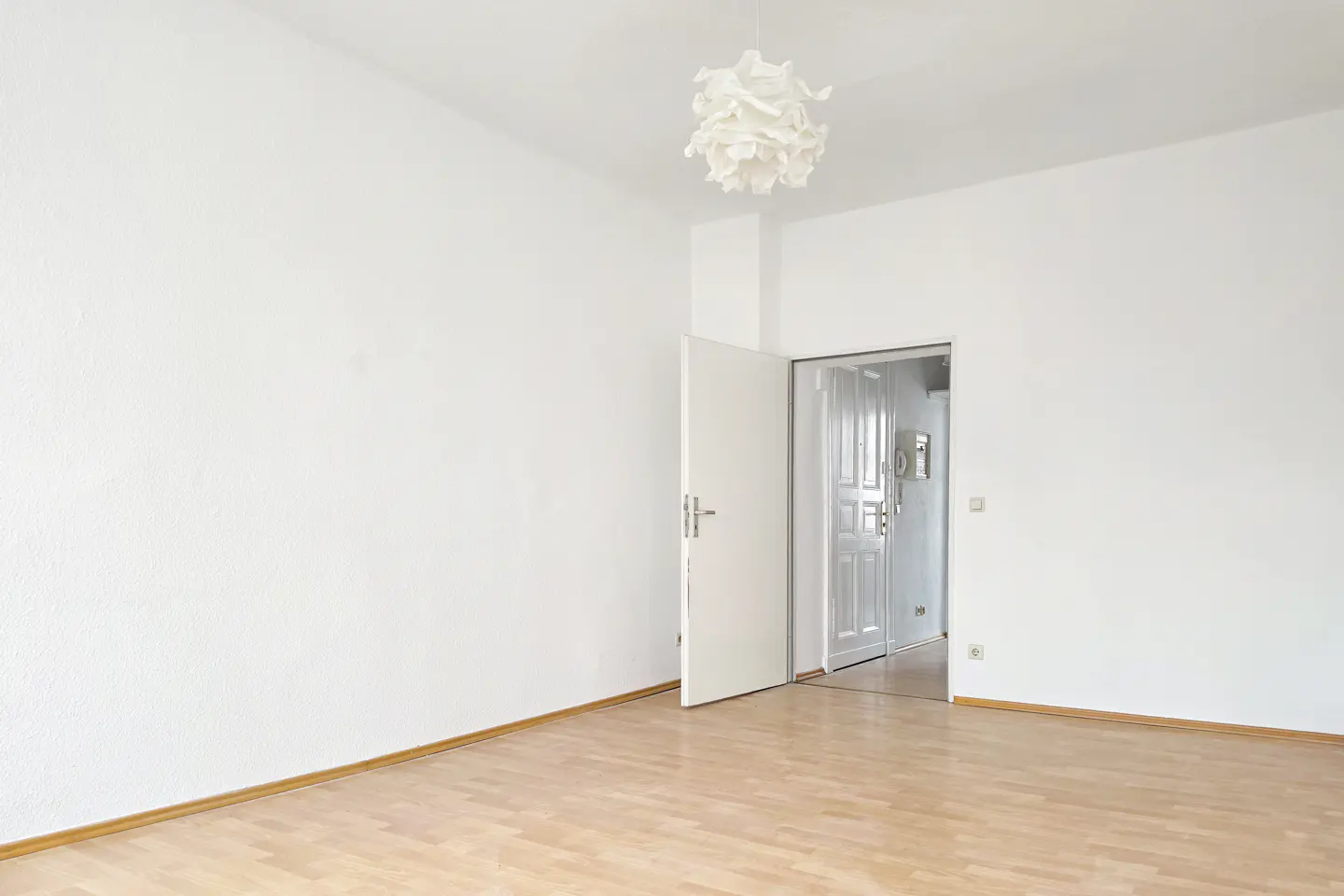 Bright, empty room with white walls, wood floor, and open white door. A modern white pendant lamp hangs from the ceiling.