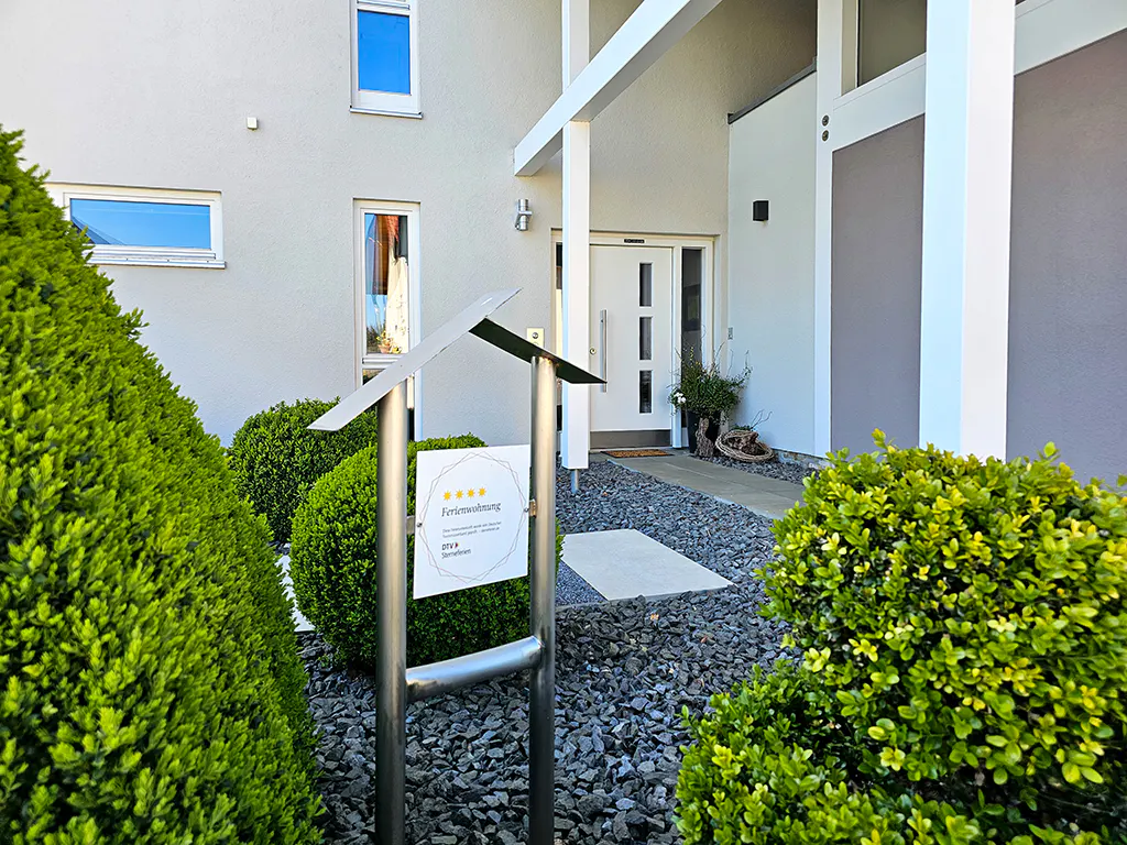 Modern home exterior with a white door, gray gravel, and green bushes. A sign displays "Ferienwohnung" with five stars.