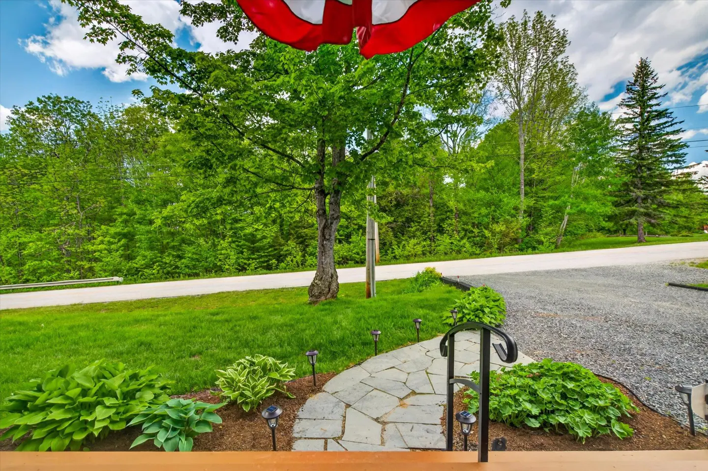 A stone path leads through a green lawn with lush plants and a large tree, viewed from a porch under a red umbrella.