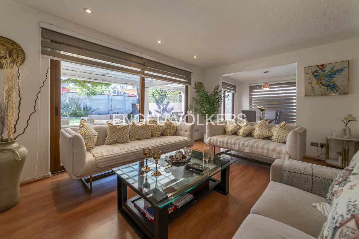 Living room with two beige sofas, patterned pillows, glass coffee table, and wood floors. Large windows overlook a backyard.