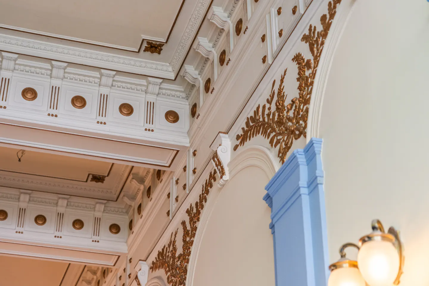Ornate white ceiling and wall with gold leaf details, blue column, and a blurred light fixture.