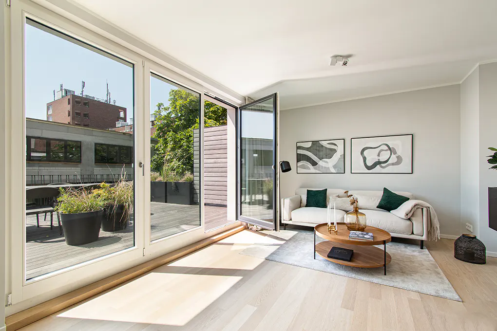 Bright living room with white sofa, wood coffee table, and abstract art. Large glass doors open to a deck with plants and city views.