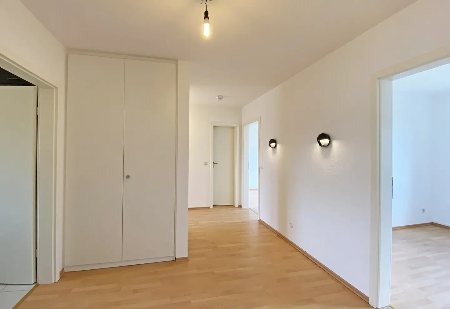 Bright hallway with light wood floors, white walls, and built-in white closet. Black sconces and a single pendant light illuminate the space.