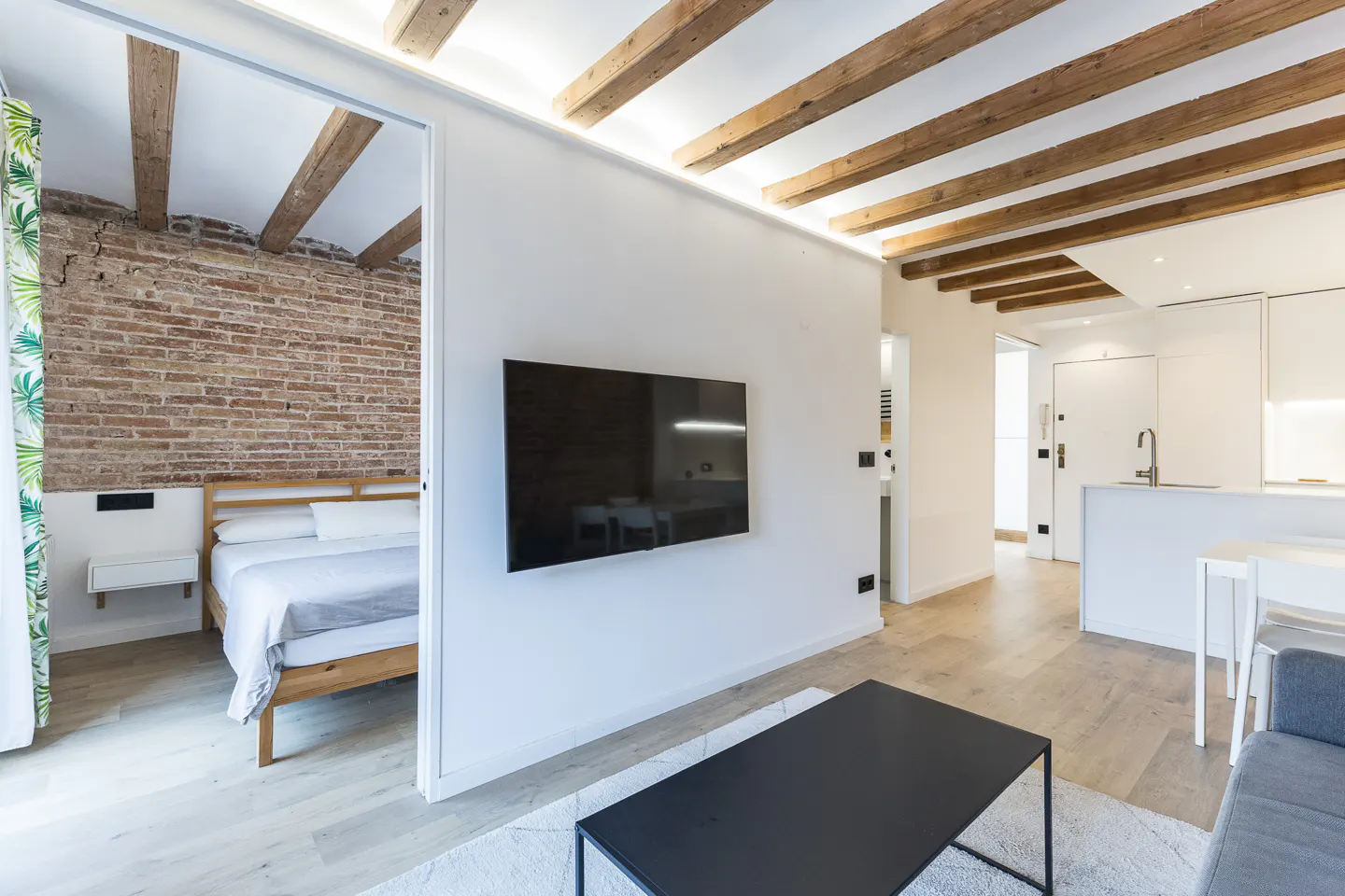 Modern apartment interior with exposed wooden beams, brick wall, and a flat-screen TV. The color palette is mostly white and neutral.