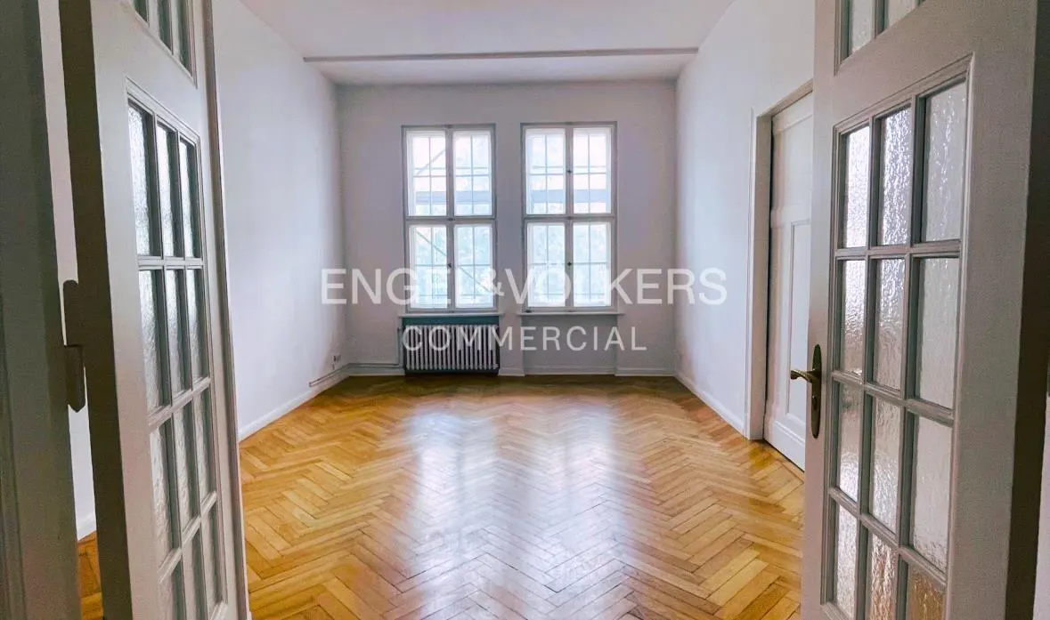 A bright, empty room with herringbone wood floors, viewed through open white doors. Two windows and a radiator are visible.
