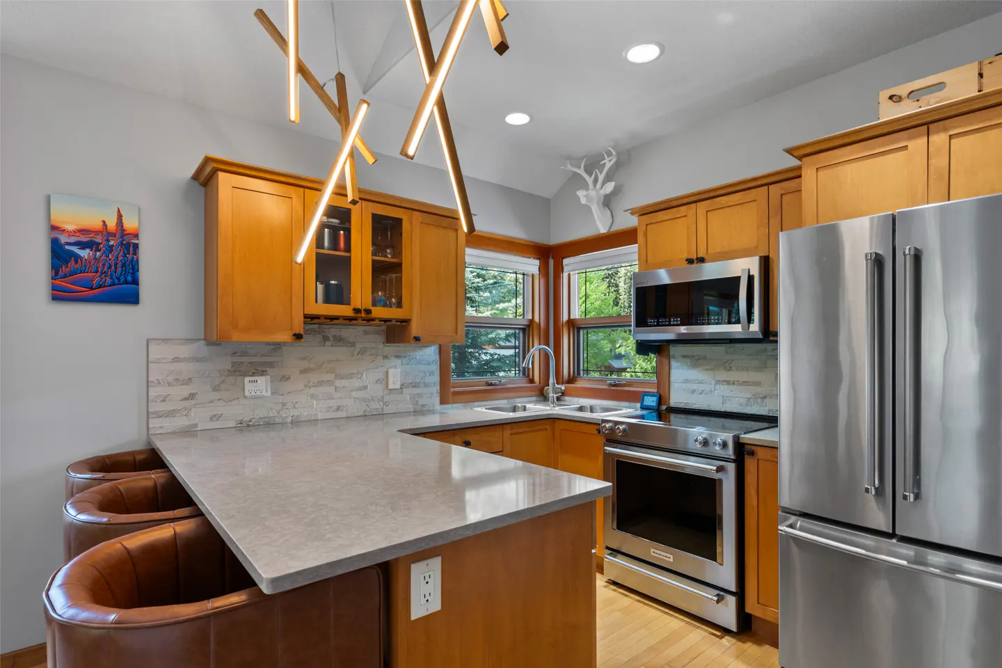 A modern kitchen with wood cabinets, stainless steel appliances, and a gray countertop with three brown leather bar stools. A painting hangs on the gray wall.