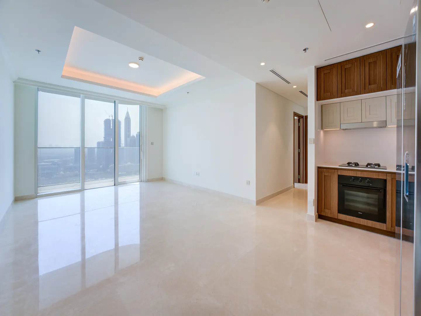 Bright, empty apartment with a city view. Large windows, white walls, and a modern kitchen with wood cabinets.