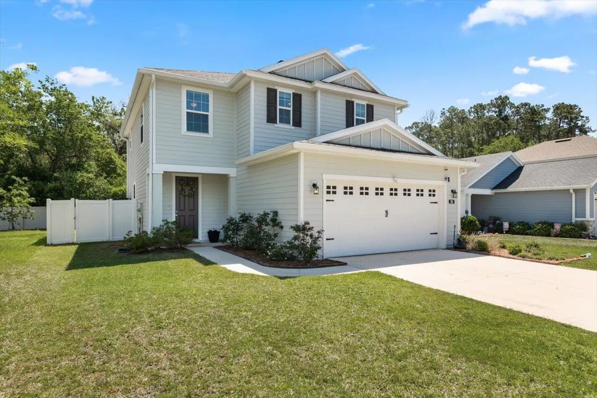 Two-story light blue house with a white garage door and black shutters, surrounded by a green lawn and trees.