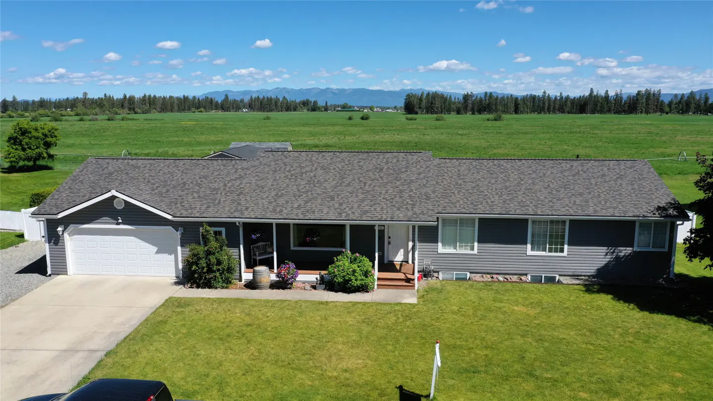 A gray ranch-style house with a green lawn, set against a backdrop of green fields and distant mountains.