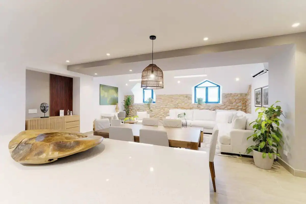 Open-concept living space with white walls, stone accent wall, wood dining table, and white sofa. A woven pendant light hangs above.