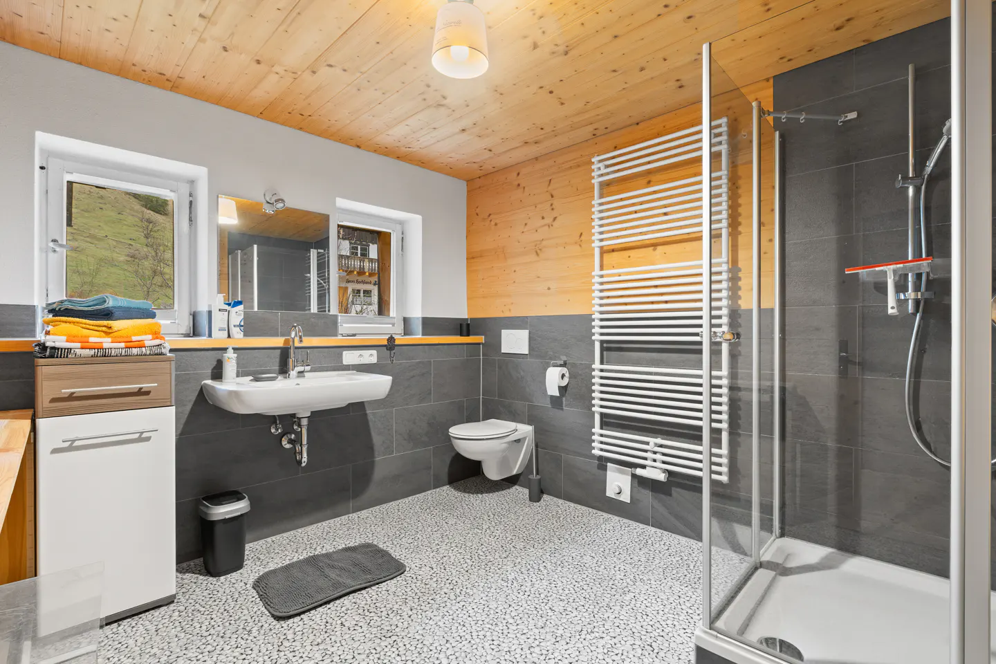 Bathroom with gray tile, wood ceiling, and pebble floor. Sink, toilet, towel warmer, and glass shower are visible.