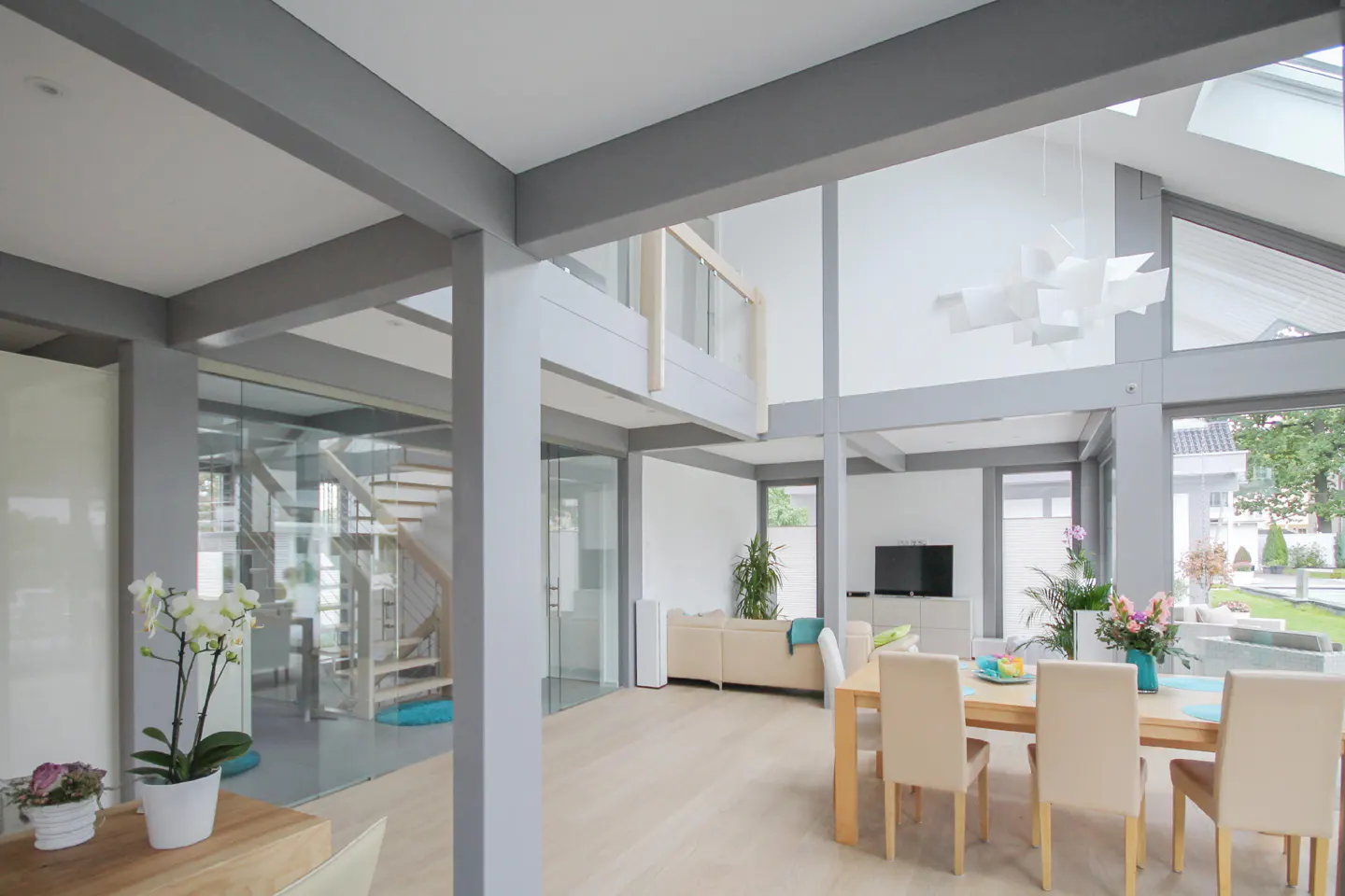 Modern open-plan living area with high ceilings, grey structural beams, a dining set, and a glass-enclosed staircase.