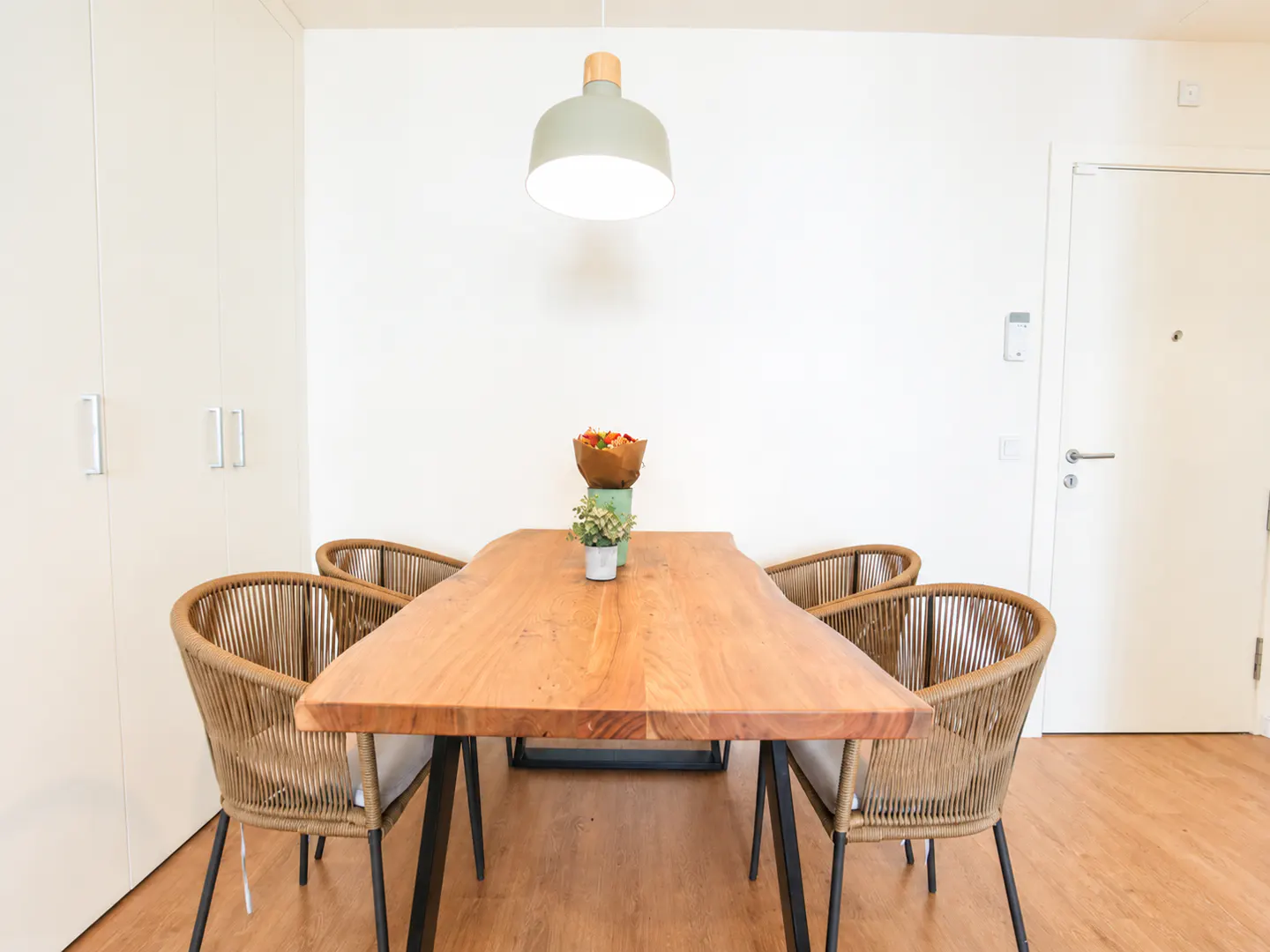 Dining area with a wood table, four wicker chairs, and a green pendant light. A bouquet and plant decorate the table. White walls and door.