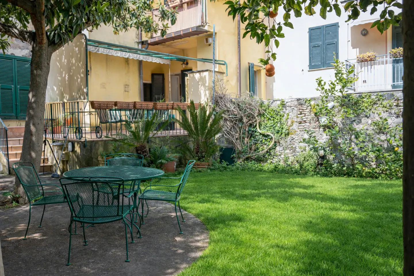 A backyard with a green metal table and chairs on a concrete patio, a green lawn, and a stone wall with ivy.