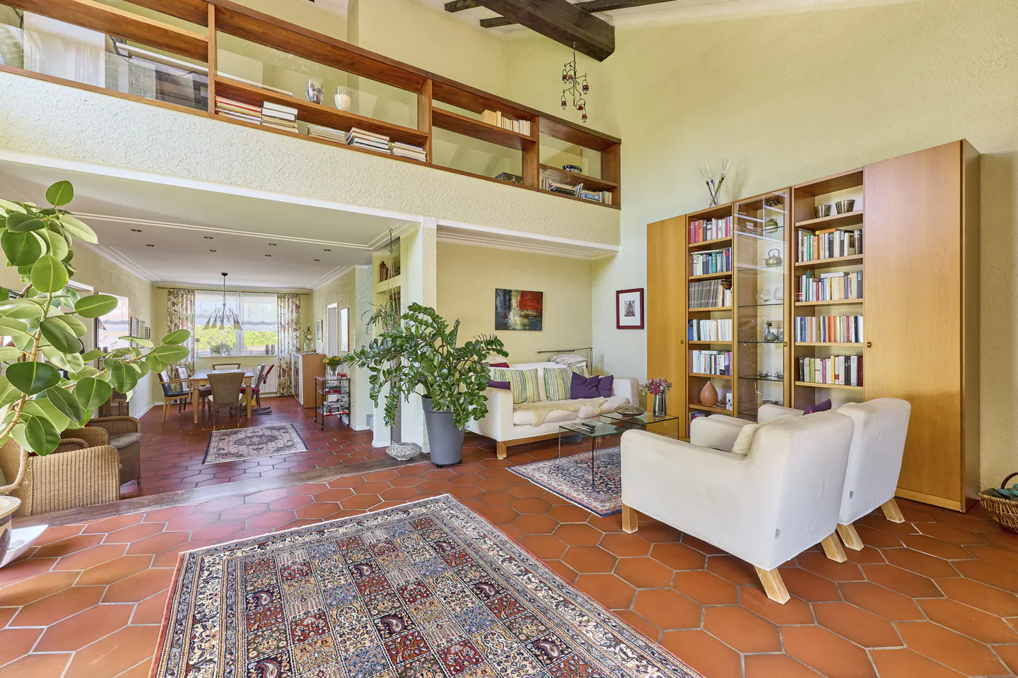 A bright living room with terracotta floors, white sofas, bookshelves, and a dining area in the background.