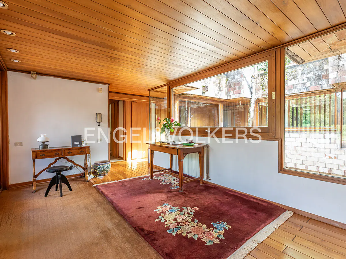 A bright room with wood ceilings, a large window, and a floral rug. A wooden table with flowers sits near the window.