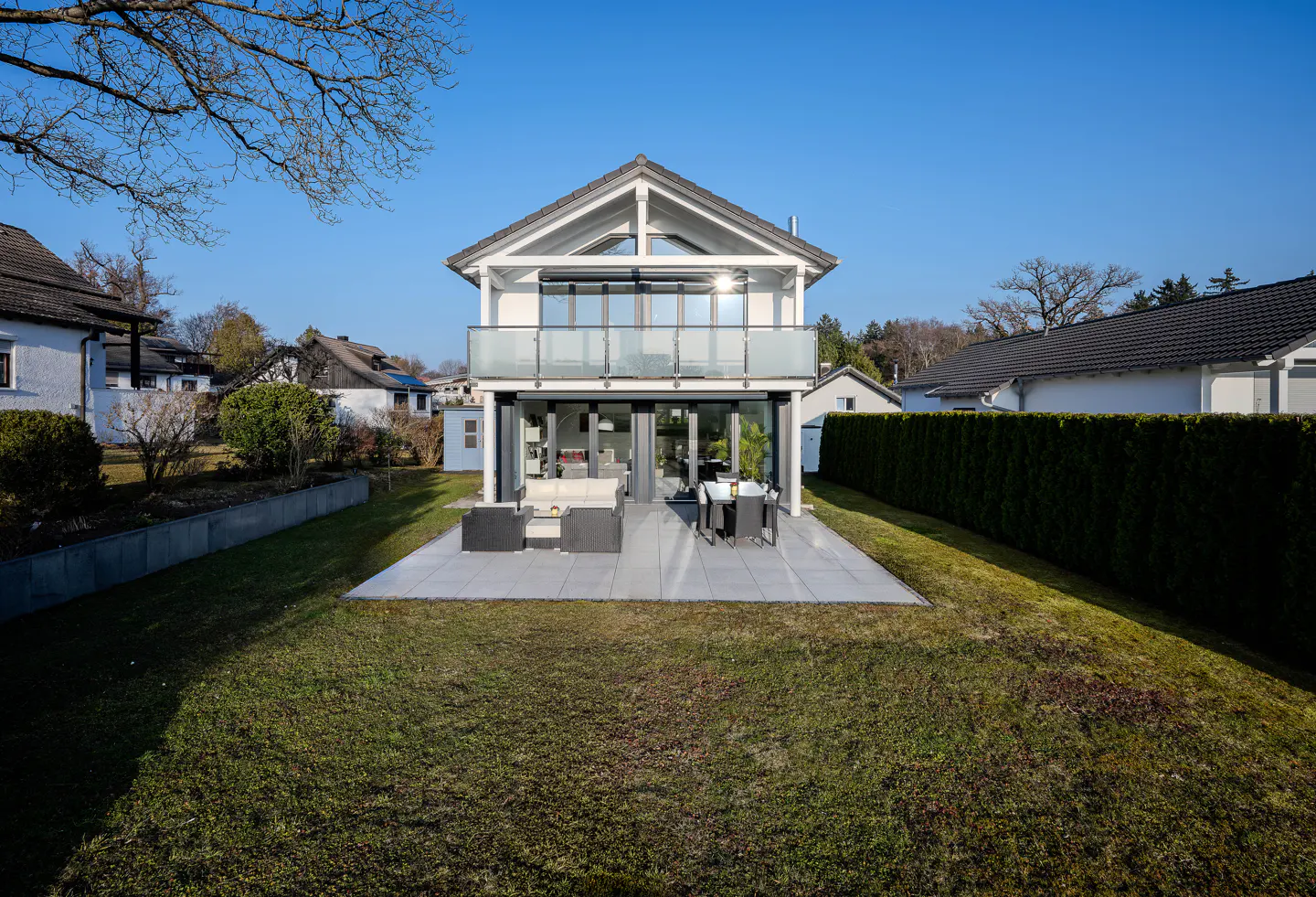 Two-story white house with a balcony and a gray patio with outdoor furniture, surrounded by a green lawn and trees.