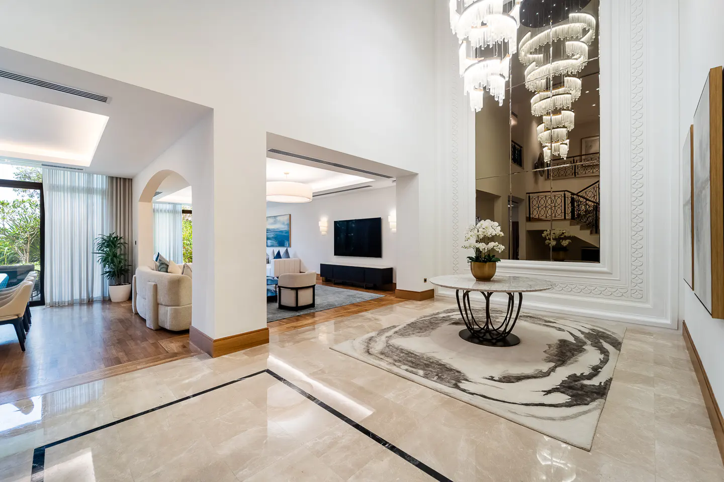 A bright foyer with marble floors, a round table with flowers, and a large mirror reflecting a staircase and chandelier.
