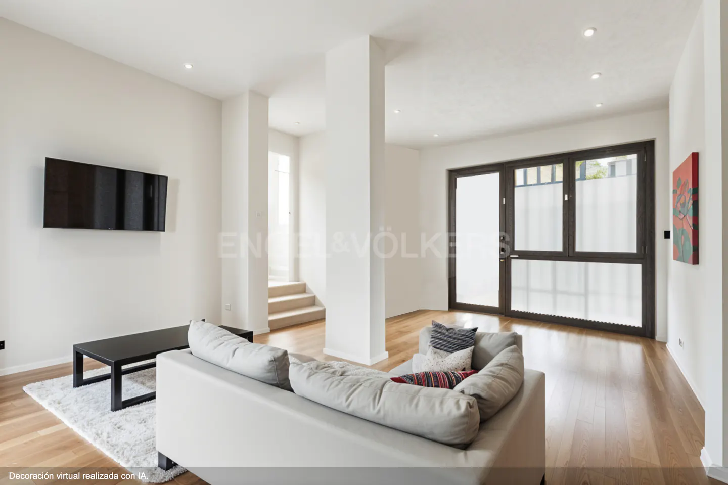 Bright living room with hardwood floors, white walls, and a gray sofa. A black TV hangs above a black coffee table on a white rug. Stairs are visible in the background.
