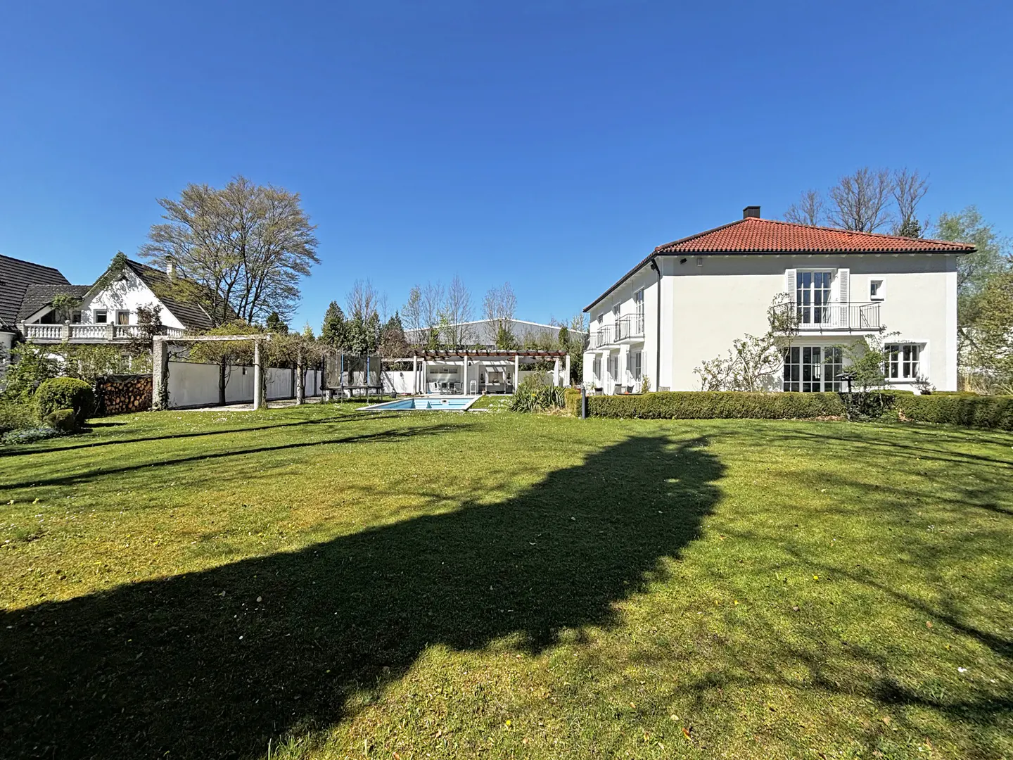 A white two-story house with a red tile roof overlooks a green lawn and pool on a sunny day.